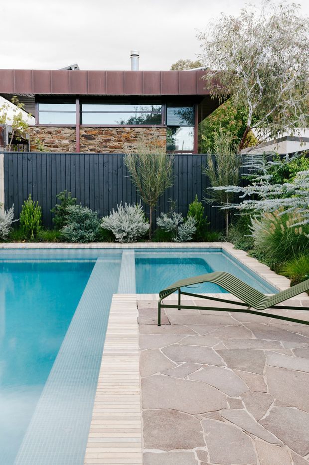 A landscaped residential pool area with a rectangular turquoise swimming pool, pale timber pool edging, irregular natural stone paving, and a green sculptural lounge chair. Behind the pool is a dark vertical timber fence, layered planting, and a modern single-storey home with rust-toned metal roof cladding, long horizontal glazing, and a natural stone wall.