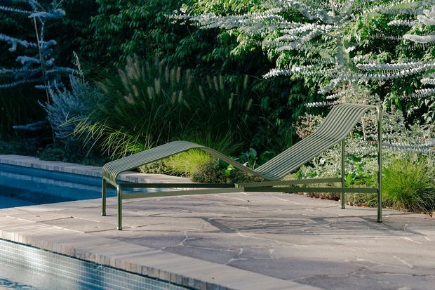 A sculptural ribbed outdoor chaise lounge with a green metal frame sits on a natural stone-paved pool deck beside a swimming pool, surrounded by dense ornamental planting and trees.