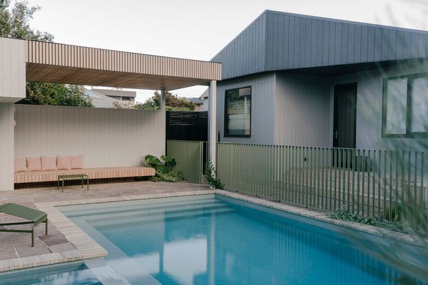 A contemporary residential courtyard viewed from beside a swimming pool. The house features dark grey-blue vertical cladding, a pitched roof form, large dark-framed glazing, and a matching covered outdoor structure with vertical slatted screening. A paved pool terrace includes built-in bench seating, lounge furniture, and planted edges.