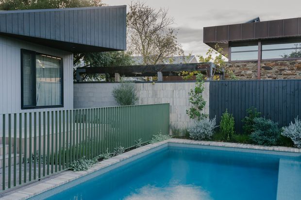 Contemporary residential courtyard with a turquoise swimming pool, dark vertical metal fencing, a pale rendered masonry wall, and angular pavilion-like building forms. The scene includes a low stone-clad wall, planted borders, and restrained blue-grey and green tones under an overcast sky.