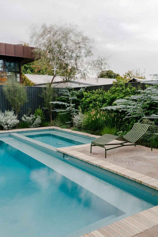 A landscaped residential pool area with a turquoise tiled swimming pool, pale stone paving, and timber decking. Two green metal-framed lounge chairs sit beside dense ornamental planting and dark vertical fencing. A modern home and neighbouring rooflines are partially visible beyond the garden.