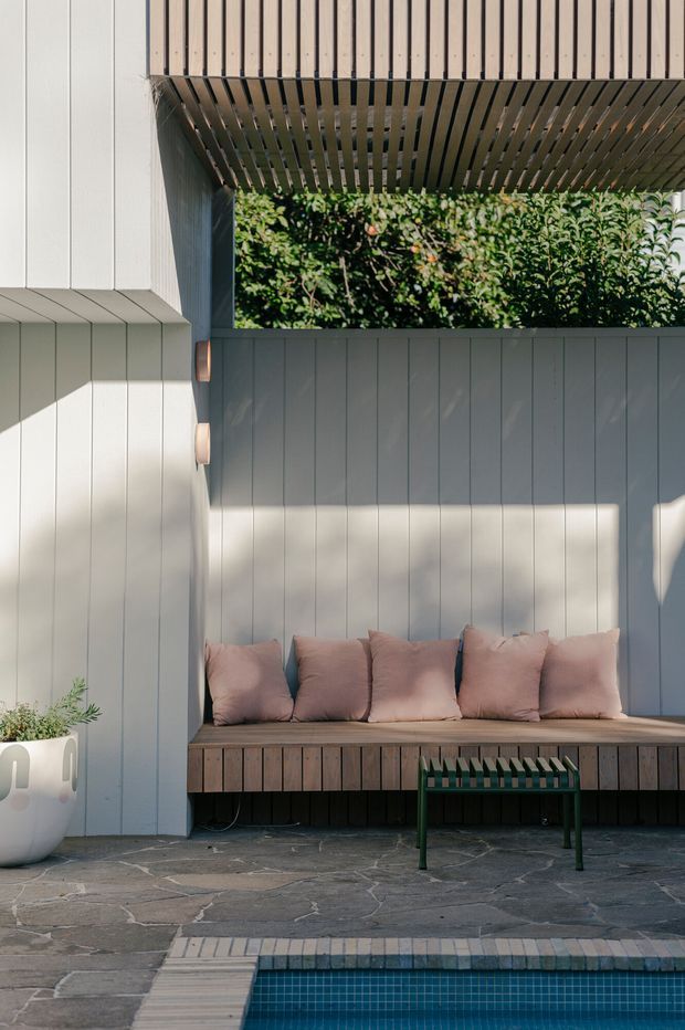 A refined outdoor poolside lounge nook with a built-in timber bench against pale vertical wall panelling. Soft blush cushions, a slatted timber pergola overhead, garden greenery, stone paving, and the edge of a tiled swimming pool create a calm, sheltered courtyard setting.
