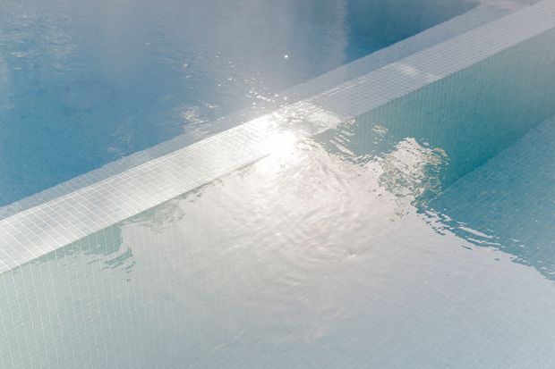 Close-up view of a swimming pool corner and water surface, featuring pale blue mosaic pool tiles, a tiled waterline or dividing edge, and bright sunlight reflecting across the rippling water. No surrounding architecture or furnishings are visible.