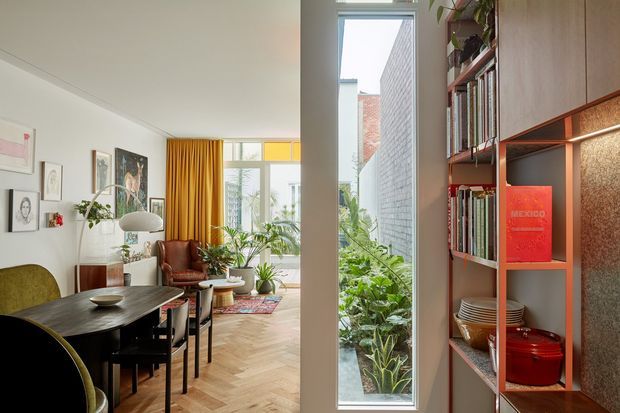 Bright, eclectic residential interior showing a dining area opening into a plant-filled living space, with herringbone timber flooring, dark dining furniture, artwork, mustard curtains, and a built-in open shelving and cabinetry unit beside a tall window. The shelving displays books, ceramics, and cookware, while a narrow glazed opening looks onto a planted courtyard or lightwell.