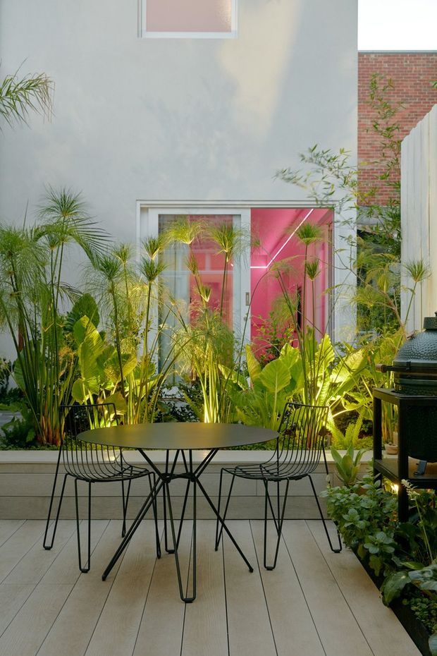 A lush courtyard-style outdoor dining area with a round black metal table and wire-frame chairs set on a pale tiled terrace. Dense tropical planting surrounds the space, with a light rendered wall, bright pink glazed doors or windows, and a red brick wall visible beyond. A dark built-in or freestanding barbecue occupies the right side.