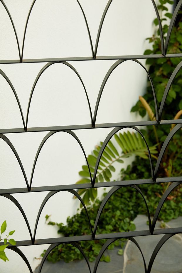 Close-up view of a repeating black metal arched screen or grille, with a white wall and lush green planting visible behind it. The geometric pattern creates a decorative architectural partition or facade element.
