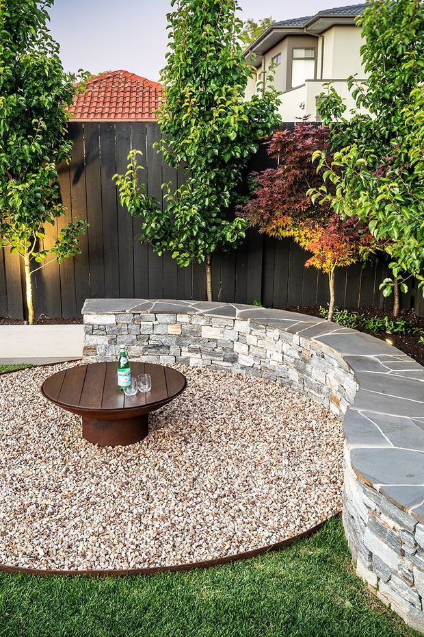 A landscaped residential courtyard or backyard with a curved stacked-stone retaining wall and dark stone cap, a circular pebble-covered sitting area, and a low round timber outdoor table. Dense ornamental trees and Japanese maple plantings sit against a dark timber privacy fence, with a neighbouring two-storey house visible beyond.