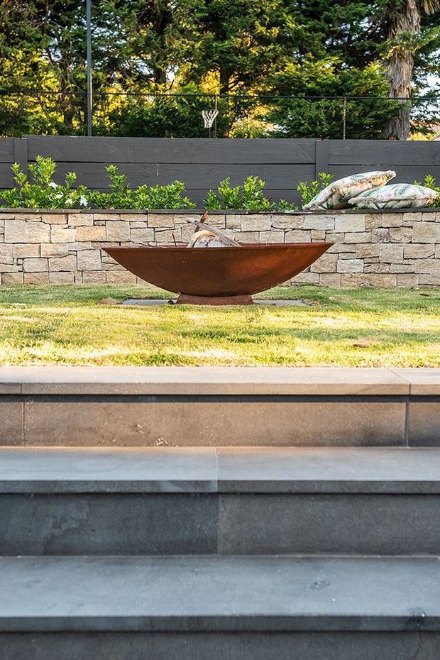 A landscaped outdoor area featuring broad dark stone steps in the foreground, a raised lawn and a low stacked-stone retaining wall. A rusted corten-steel bowl-shaped fire pit or water feature sits centrally on the grass, with planting and a dark privacy fence behind it.