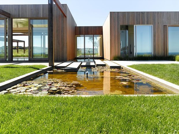 A low, modern residence arranged around a central reflecting pond with lily pads and stepping stones. The building features rectilinear timber-clad volumes, extensive floor-to-ceiling glazing, dark metal framing, and a covered connecting passage. Manicured lawn surrounds the water feature, with open landscape visible through the house.