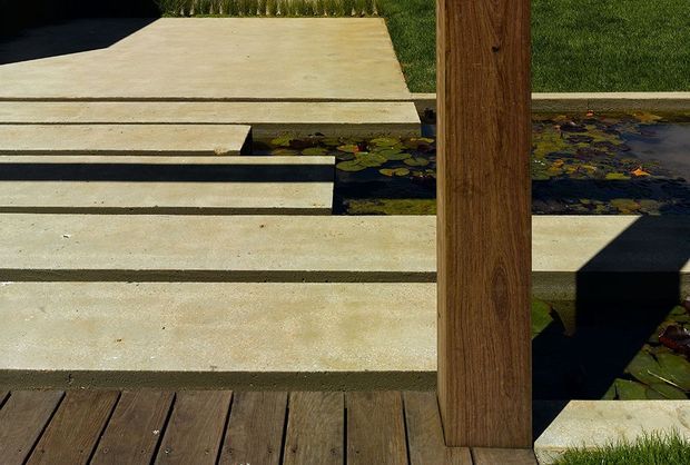 A close-up architectural landscape detail showing broad pale concrete steps and slabs spanning a shallow water feature with lily pads. A dark timber deck occupies the foreground, while a substantial vertical timber post divides the scene. Trimmed lawn and planting are visible around the hard landscaping, with strong sunlight creating sharp shadows.
