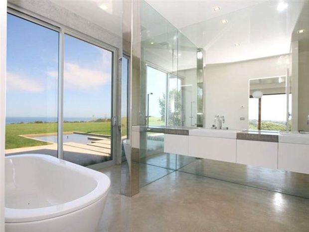 Bright contemporary bathroom with a freestanding white bathtub, expansive frameless glass shower enclosure, floating white vanity with dark stone benchtop, large mirror, polished concrete flooring, and floor-to-ceiling glazing overlooking a landscaped coastal setting and pool.
