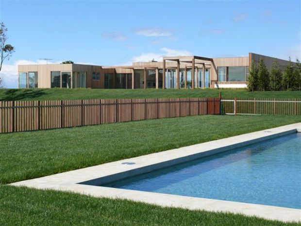 A low, contemporary single-storey residence with extensive glazing, timber-clad walls and a series of projecting framed outdoor spaces. The house sits behind a timber slat fence and landscaped lawn, with a rectangular swimming pool occupying the foreground.