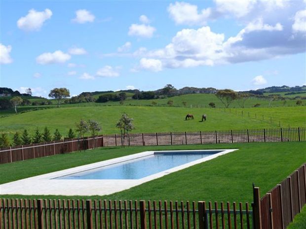 A landscaped rural property with a long rectangular swimming pool, broad manicured lawn, dark timber fencing, and open green pasture beyond. The scene is viewed outdoors under a bright blue sky with scattered clouds.