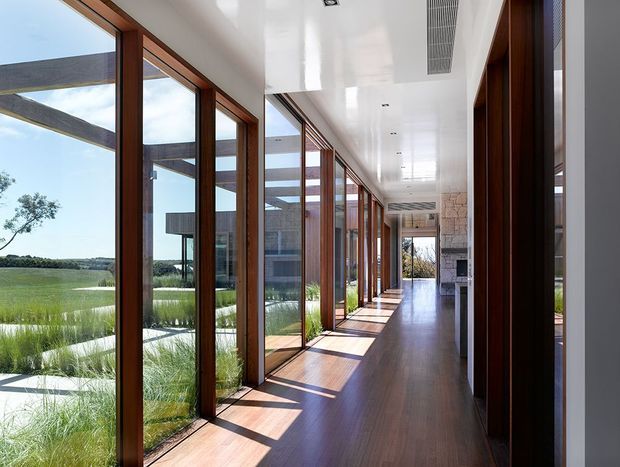 A bright, elongated interior hallway lined with full-height timber-framed glazing on one side, opening onto a landscaped outdoor area with water and grasses. The corridor has dark timber flooring, white walls and ceiling, recessed lighting, linear air-conditioning grilles, and a stone feature wall at the far end. Strong sunlight creates geometric shadows across the floor.