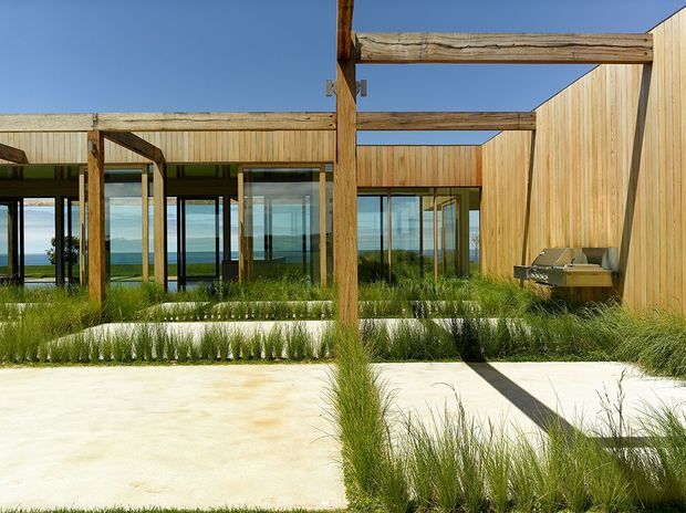 A contemporary coastal courtyard and outdoor entertaining area with pale concrete paving, native grasses, timber-clad walls, exposed timber beams and extensive floor-to-ceiling glazing. A built-in stainless-steel barbecue sits against the timber wall, while the ocean and coastal landscape are visible beyond the glass.