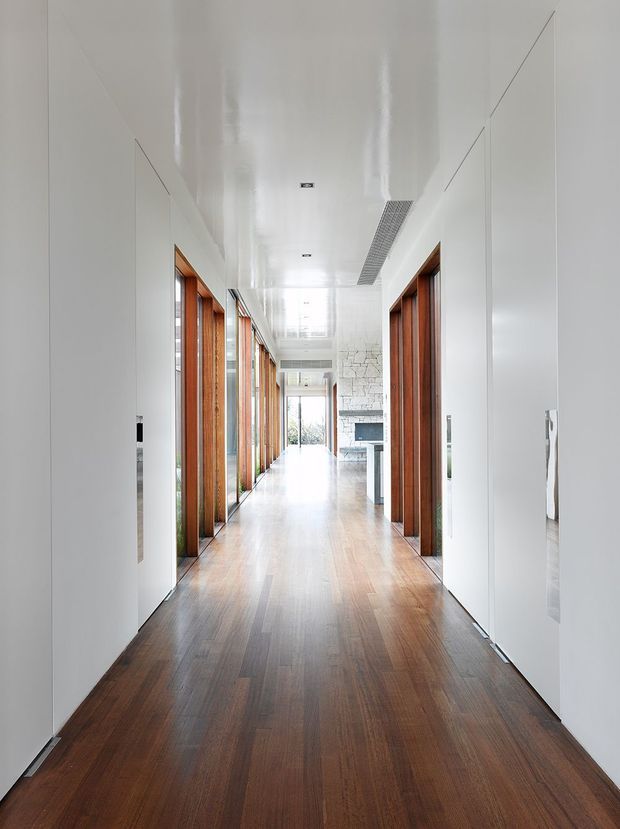 A long, bright residential hallway with rich dark timber flooring, white walls and a glossy white ceiling. Full-height timber-framed glazed openings line the left side, while timber-lined doorways and concealed white doors line the right. The corridor leads toward an open living area with a stone-clad fireplace visible at the far end. The design is clean, minimalist and strongly linear, with abundant natural light.