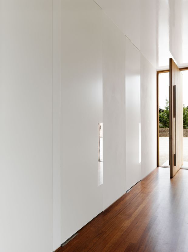 Minimal, bright interior hallway or entry space with uninterrupted white built-in wall panels, concealed openings, and warm timber flooring. A tall timber-framed glazed door opens to the exterior, bringing in strong natural light. The space is sparsely furnished and highly refined.