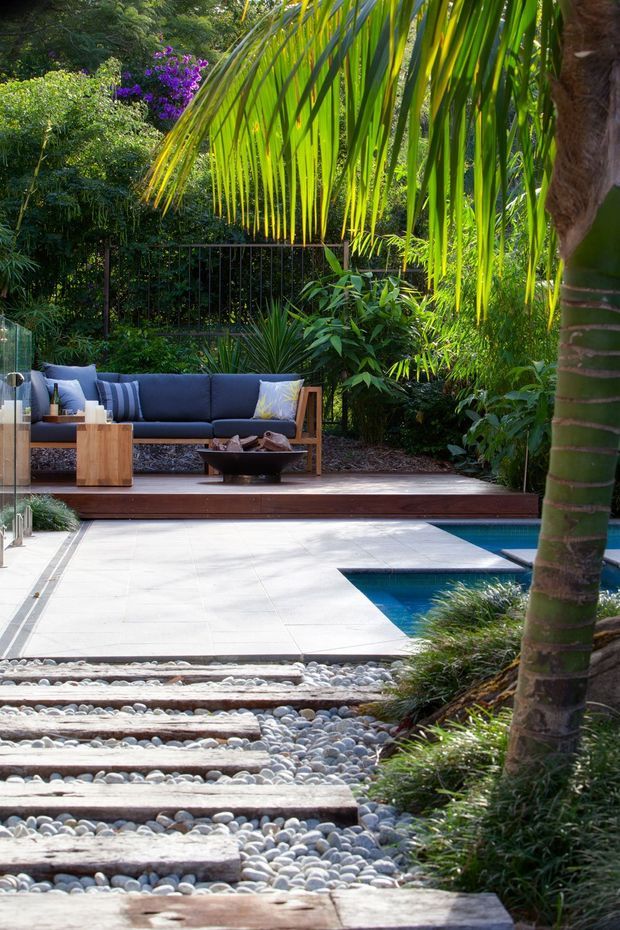 A lush tropical outdoor poolside courtyard with a timber-framed lounge setting on a raised timber deck. Large palm foliage and dense planting surround a blue swimming pool, while pale stone paving and a decorative pathway of timber sleepers and smooth river pebbles lead through the garden.