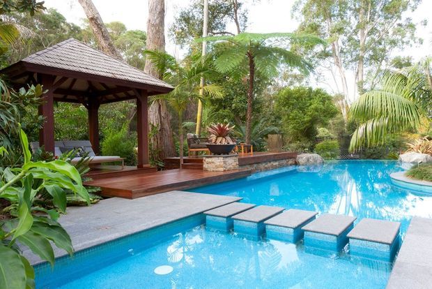 A tropical outdoor pool area surrounded by lush vegetation, featuring a bright blue swimming pool with stepping-stone pads, a timber deck, and a dark timber pavilion with a shingled pitched roof. A raised stone-edged feature and seating area sit beside the pool.