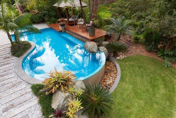 A tropical landscaped pool area with a curving bright-blue swimming pool edged in smooth concrete and natural rocks. A timber deck leads to a small roofed outdoor pavilion furnished with seating, while lush palms and dense planting surround the pool. A pale gravel-and-paver path borders the lawn and garden.