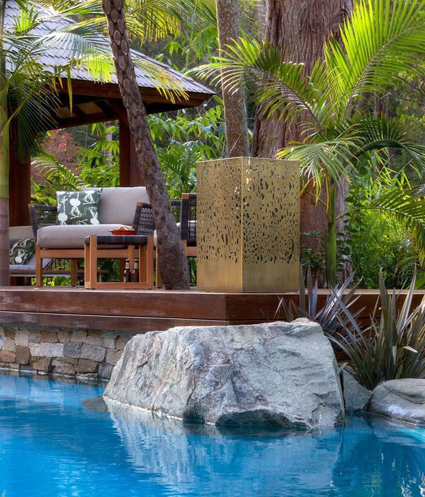 A lush tropical outdoor pool area with a raised timber deck, natural stone retaining wall, large boulders integrated into the pool edge, and dense palm planting. The deck includes cushioned outdoor seating beneath a timber-framed roof structure and a perforated brass-toned privacy screen or feature panel. The scene has a resort-like, organic character with strong natural materials and vibrant blue water.