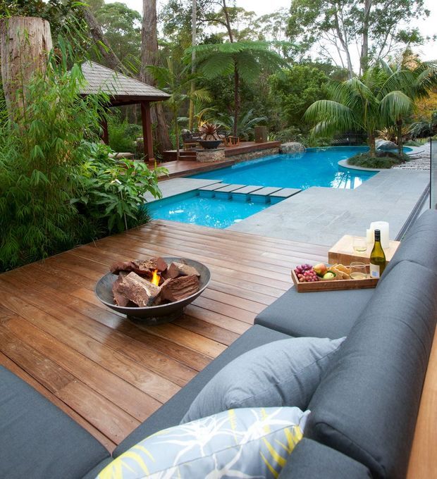 A lush tropical outdoor entertaining area with a timber pool deck, bright blue swimming pool, integrated stepping platforms, and a covered pavilion with a pitched roof. A large outdoor sectional sofa, fire bowl, and drinks create a relaxed resort-style setting surrounded by dense greenery and palms.