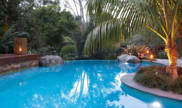 A tranquil landscaped swimming pool surrounded by dense tropical planting, large natural boulders, palms, and warm outdoor lighting. A low stone-clad feature wall with an illuminated vertical element borders the pool, creating a resort-like garden setting.