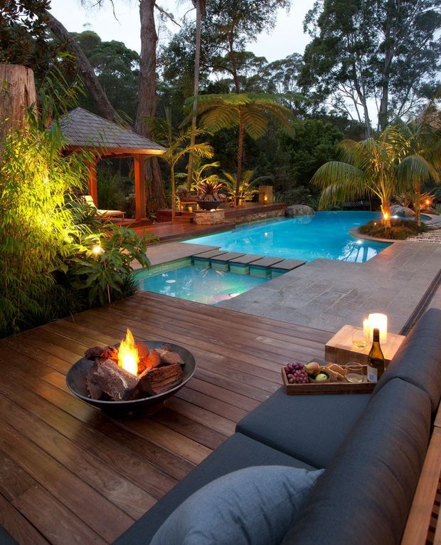 A lush tropical landscaped pool setting viewed from an outdoor timber deck. The deck features a built-in dark upholstered lounge, a fire bowl with burning logs, side table with drinks and fruit, and warm landscape lighting. A turquoise swimming pool with integrated shallow spa and stepping stones occupies the centre, with a small timber pavilion and dense mature greenery surrounding the space.