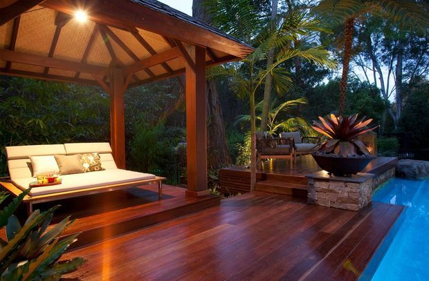 A lush tropical outdoor entertaining area with a timber deck beside a swimming pool. The space features a covered timber pavilion with exposed rafters and posts, built-in cushioned seating, timber steps, stone-clad planter or seat walls, and dense tropical planting. Warm integrated lighting creates an inviting resort-like atmosphere.