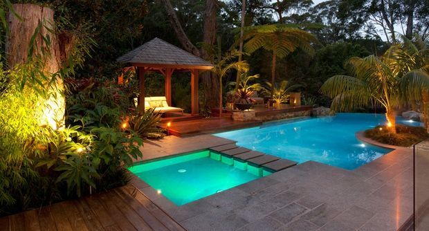 A tropical landscaped pool area at dusk, featuring a turquoise swimming pool with an integrated illuminated spa, dark stone paving, timber decking, lush palms and dense planting, and a timber-roofed poolside pavilion with seating. Warm landscape lighting creates a resort-like atmosphere.