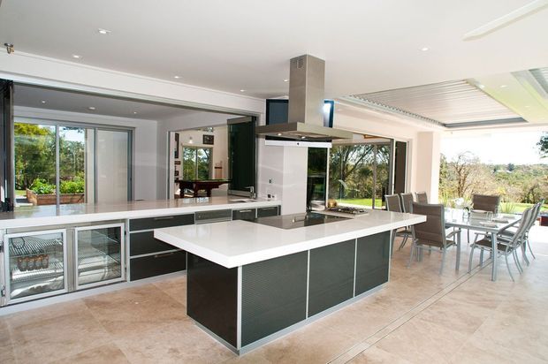 Bright, spacious open-plan kitchen and dining area with a large white island, dark built-in cabinetry, integrated wine and beverage refrigerators, stainless-steel rangehood, and expansive glazed doors opening to a leafy outdoor setting. A dining table sits beside the kitchen, with pale tiled flooring and a clean contemporary fit-out.