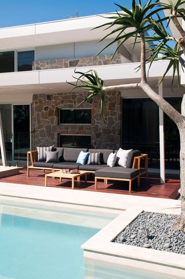 A contemporary two-storey residence with crisp white horizontal forms, extensive dark-framed glazing, and prominent irregular natural-stone feature walls. A timber outdoor deck fronts a large dark grey sectional seating area and coffee table beside a turquoise swimming pool. Sculptural tropical trees and a gravel garden bed complete the sunlit outdoor setting.