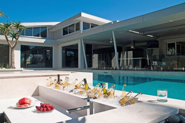 A bright contemporary residential courtyard with a turquoise swimming pool, large glazed openings, and a covered outdoor entertaining area. In the foreground, a white stone outdoor bar or kitchen island incorporates an ice trough filled with champagne bottles, with fruit plates and a drink arranged on the counter. The home features clean horizontal lines, flat roofs, white rendered surfaces, dark window frames, and integrated outdoor living spaces.