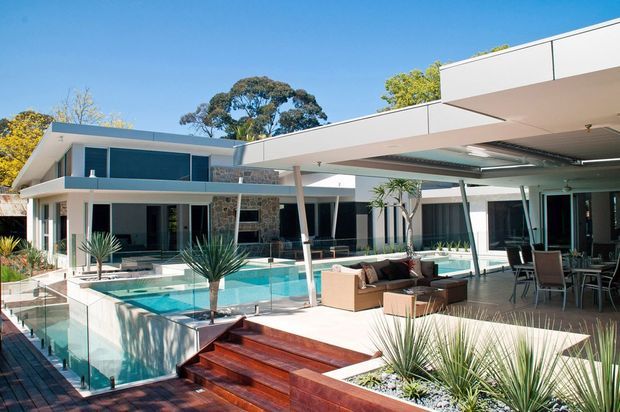 Modern single-storey residence with broad flat roof planes, extensive glazing, and stone feature walls arranged around a bright rectangular swimming pool. The outdoor entertaining area includes a covered lounge and dining space, timber decking and steps, glass pool fencing, and drought-tolerant landscaped planting.