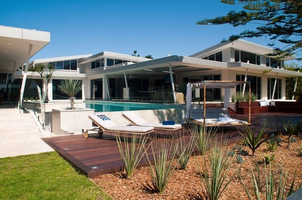 Exterior view of a contemporary flat-roofed residence with extensive glazing, white rendered forms, a covered outdoor entertaining area, and a swimming pool adjoining a timber deck. Drought-tolerant landscaping, sun loungers, and a poolside cabana frame the outdoor setting.