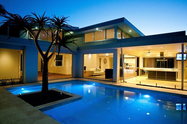Twilight exterior view of a contemporary two-storey residence arranged around a brightly illuminated swimming pool and courtyard. The home has clean horizontal rooflines, extensive glazing, a covered outdoor entertaining area, and a visible open-plan kitchen and living space. A sculptural multi-trunk planting feature sits within the pool courtyard.