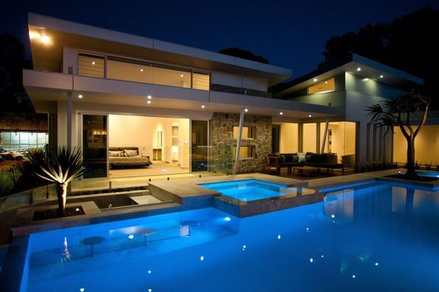 Night-time exterior view of a large contemporary multi-level residence with broad flat rooflines, extensive glazing, warm recessed lighting, a stone feature wall, and a generous illuminated swimming pool and spa in the foreground. The image is photographed from the outdoor pool area, with interior living spaces visible through the glass.
