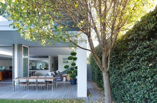 Contemporary open-plan home viewed from a landscaped outdoor deck. The scene features a clean white and dark-grey building facade, expansive glazed openings, a timber-look deck with outdoor dining furniture, and a light-filled interior living and dining area beyond. Mature trees and dense hedging frame the dwelling.
