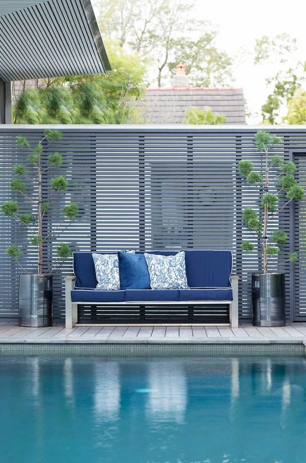 A stylish outdoor poolside seating area with a deep blue upholstered bench, patterned cushions, tall potted topiary plants, and a dark horizontal slatted privacy screen. The pool occupies the foreground, while a modern roof overhang and leafy vegetation frame the scene.