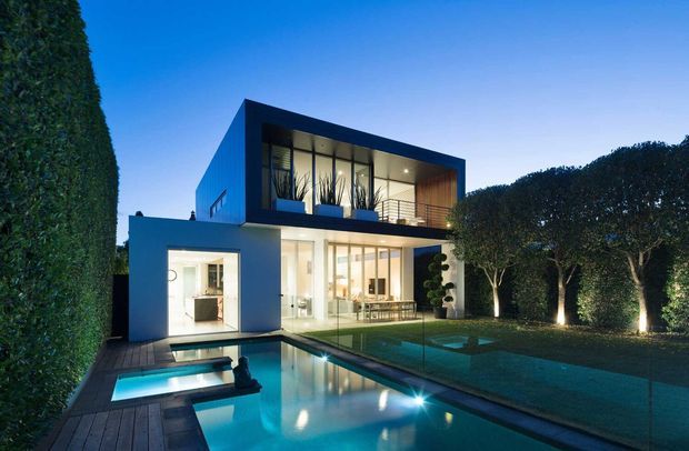Twilight exterior view of a contemporary two-storey flat-roof residence with strong geometric volumes, extensive floor-to-ceiling glazing, and a cantilevered upper level. The home opens onto a large illuminated swimming pool and timber deck, enclosed by tall manicured hedges and landscaped garden edges.
