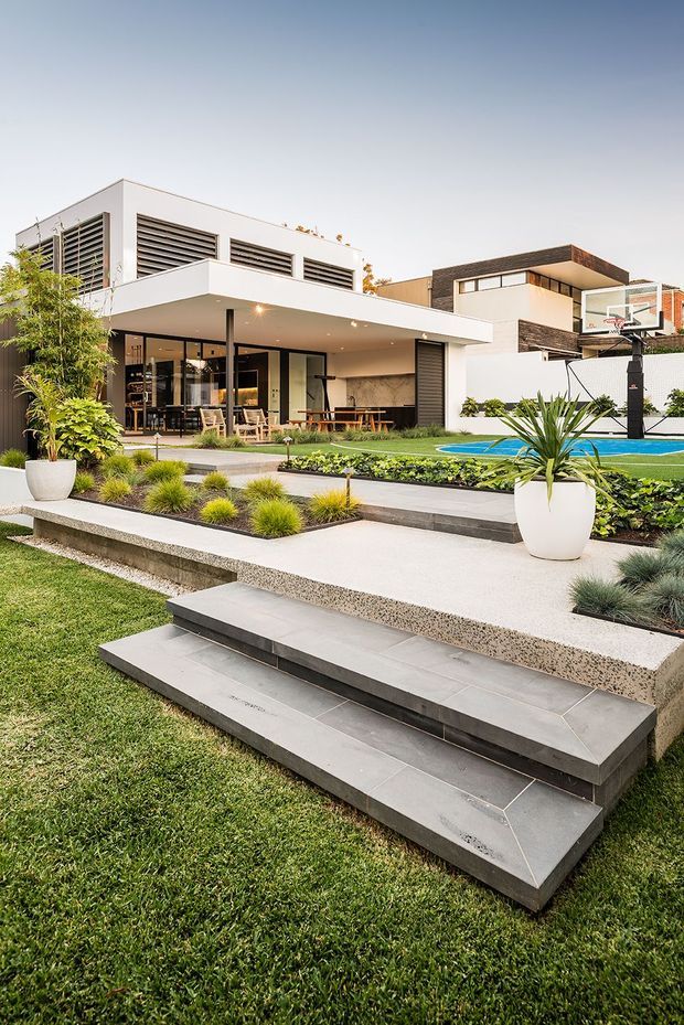 A landscaped backyard of a contemporary multi-level home, featuring broad tiled steps, concrete pathways, manicured lawn and planting beds. The house has a flat-roofed white facade with extensive glazing, horizontal louvres, and a covered outdoor entertaining area. A bright blue swimming pool and basketball hoop are visible beside the patio.