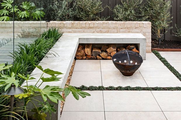 A landscaped outdoor courtyard or patio featuring a built-in light stone fireplace with stacked firewood, large pale square pavers, lush planting, olive trees, and a freestanding dark metal fire bowl. A dark vertical slatted fence forms the backdrop, creating a contemporary outdoor entertaining setting.