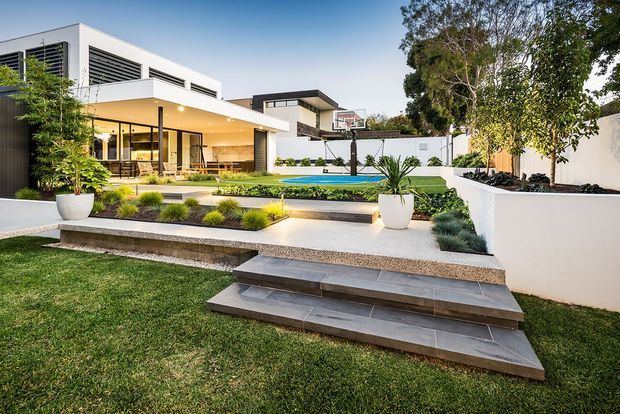 A contemporary two-storey residence viewed from a landscaped front or rear garden. The home features crisp white rendered walls, broad glazed openings, dark horizontal louvres, and a covered outdoor entertaining area. Layered stone-paved steps lead through manicured lawn and planting toward a raised terrace and a narrow swimming pool, with modern raised garden beds and feature planters.