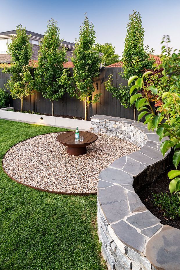 A landscaped residential backyard with a curved dry-stone retaining wall and dark stone coping, a rounded gravel seating area with a low circular timber table, lush lawn, mature ornamental trees, and a tall timber privacy fence. Suburban houses are visible beyond the fence.