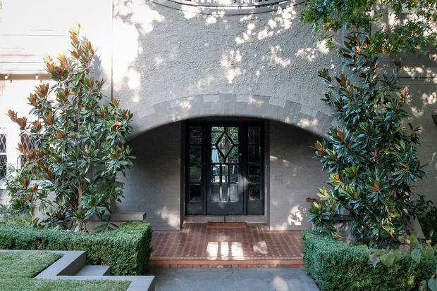 Front entrance of a refined traditional residence, featuring a textured light-grey stucco facade, a broad arched opening, dark glazed timber entry doors, and a brick-paved porch approached through formal clipped hedges and magnolia plantings.