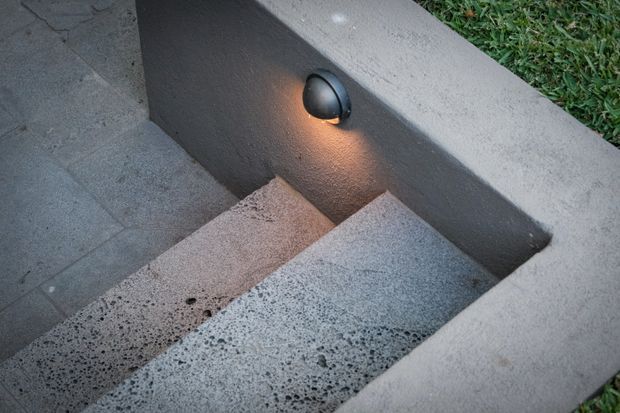 Close-up view of a short exterior stairway with broad grey textured concrete or stone steps, bordered by low rendered walls. A small wall-mounted light illuminates the stair treads, with lawn visible beyond the edge.