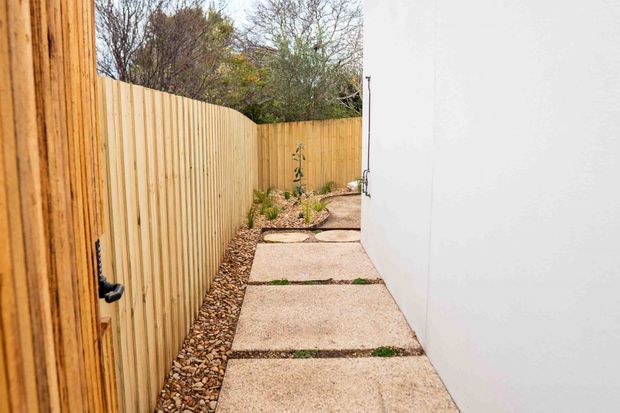 A narrow side passage with a pale concrete stepping path between a tall vertical timber paling fence and a smooth white exterior wall. Pebble landscaping borders the path, with sparse planted greenery and mature vegetation visible at the far end.