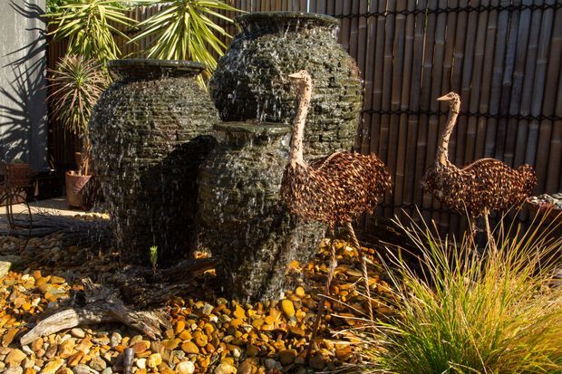A landscaped outdoor water feature with three dark textured stone urns cascading water into a pebble-covered basin. Sculptural metal bird forms, ornamental grasses, yucca-like plants, and a vertical timber privacy screen surround the fountain.