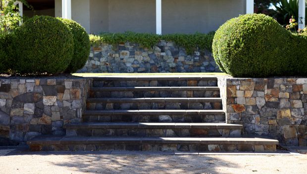 Exterior entrance steps leading up to a covered porch, framed by rounded manicured shrubs and extensive irregular natural-stone retaining walls and stair risers. The facade includes pale rendered walls and white vertical posts, with strong sunlight and landscaping visible around the entry.