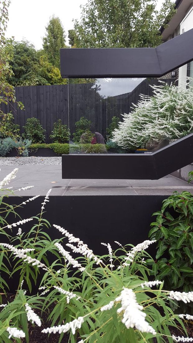 A landscaped residential courtyard or garden featuring a striking angular black architectural structure with large reflective glass panels, integrated planter-like forms, dark timber fencing, paved surfaces, and dense ornamental planting with white flowering stems. The composition emphasizes geometric hardscape and sculptural built elements set against lush greenery.